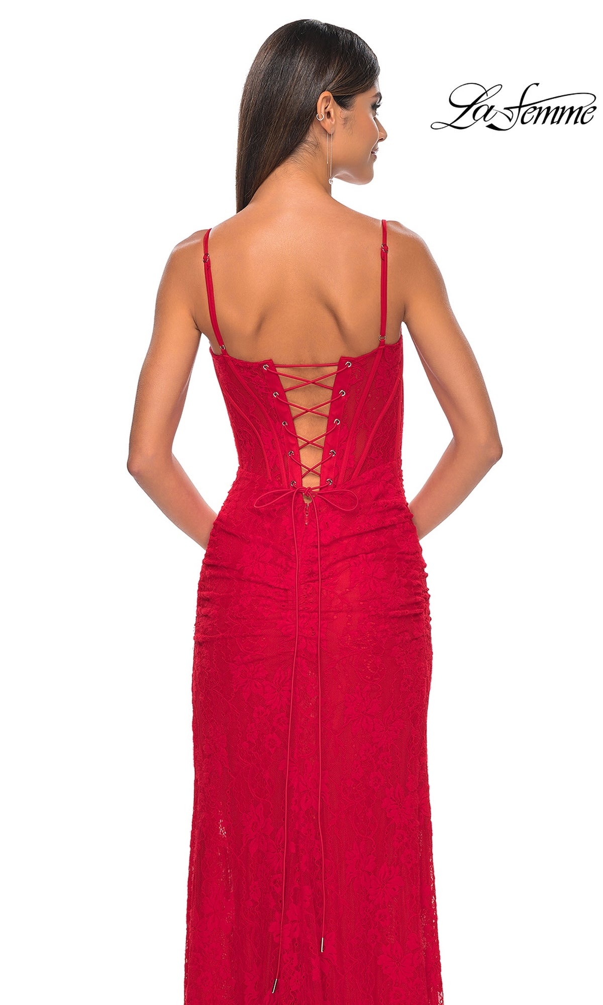 A woman stands with her back to the camera, wearing the La Femme Lace-Up Back Long Lace Prom Dress 32237, a red designer gown with thin straps and a lace-up corset-style bodice. The